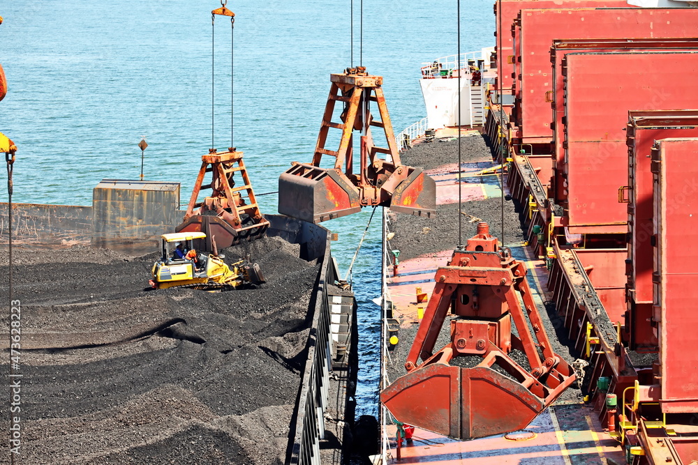 Loading coal from cargo barges onto a bulk carrier using ship cranes and grabs at the port of Muara Pantai, Indonesia. January,2021.