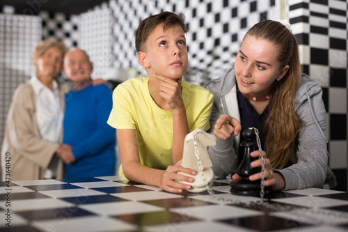 Boy and girl solving chess riddle while standing in escape room. Grandfather and grandmother standing in background and watching them.