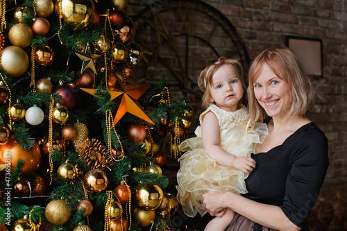 A blonde mother in a black dress and a little daughter near a Christmas tree with golden balls