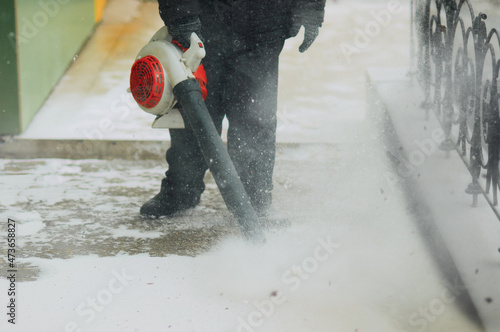 A man removes snow from the sidewalk with a wind blower. Lumps of snow and snowflakes in the air. Work outdoors in winter. Snow removal after a snowfall.
