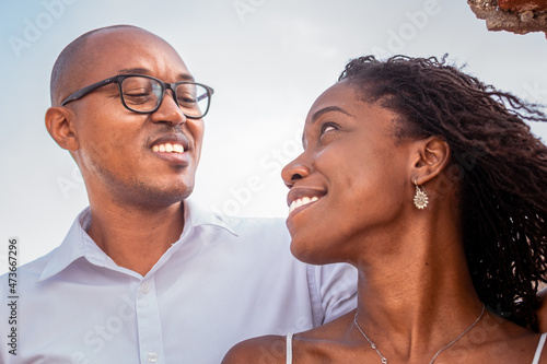 Attractive black woman smiling, looking over her shoulder at her partner. Loving African couple stare at each other, bright sky in background. Closeup looking at each other