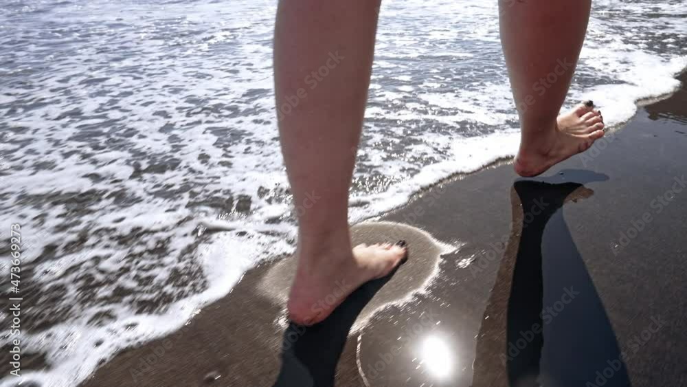 Legs of woman walking barefoot on wet black volcanic sand island beach. Beautiful feet of young girl near sea on sunset or sunrise. Slow motion. Legs of woman walking barefoot on wet black volcanic sand island beach. Beautiful feet of young girl near sea on sunset or sunrise. Slow motion.