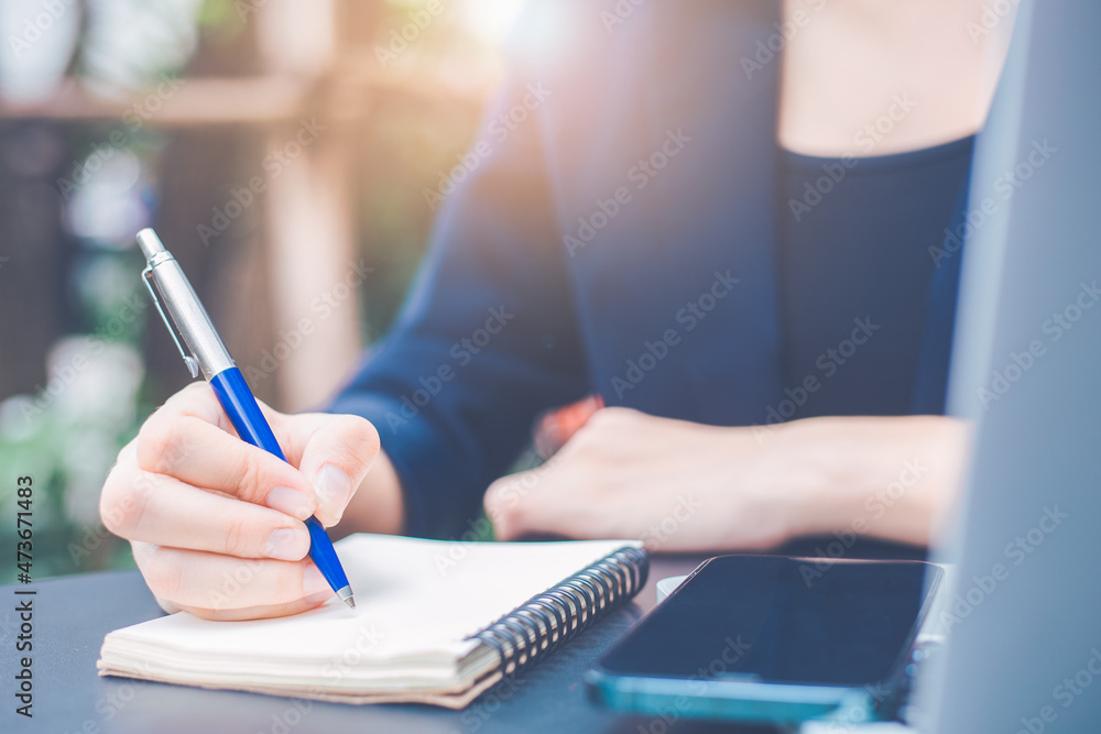 Woman hand writing on a notepad with a pen and she is holding a cell phone in the office.