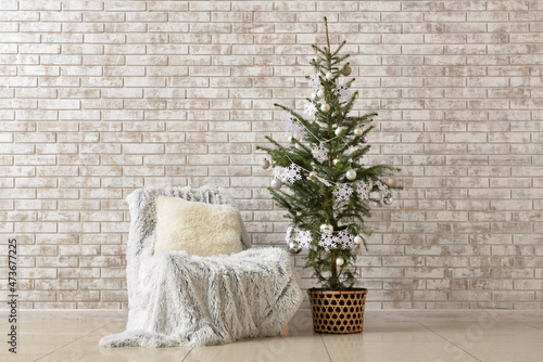 Beautiful Christmas tree in pot decorated with snowflakes, balls and comfortable armchair near beige brick wall