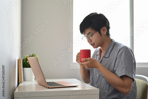 Peaceful asian man in pajamas using computer laptop in bedroom and drinking coffee.