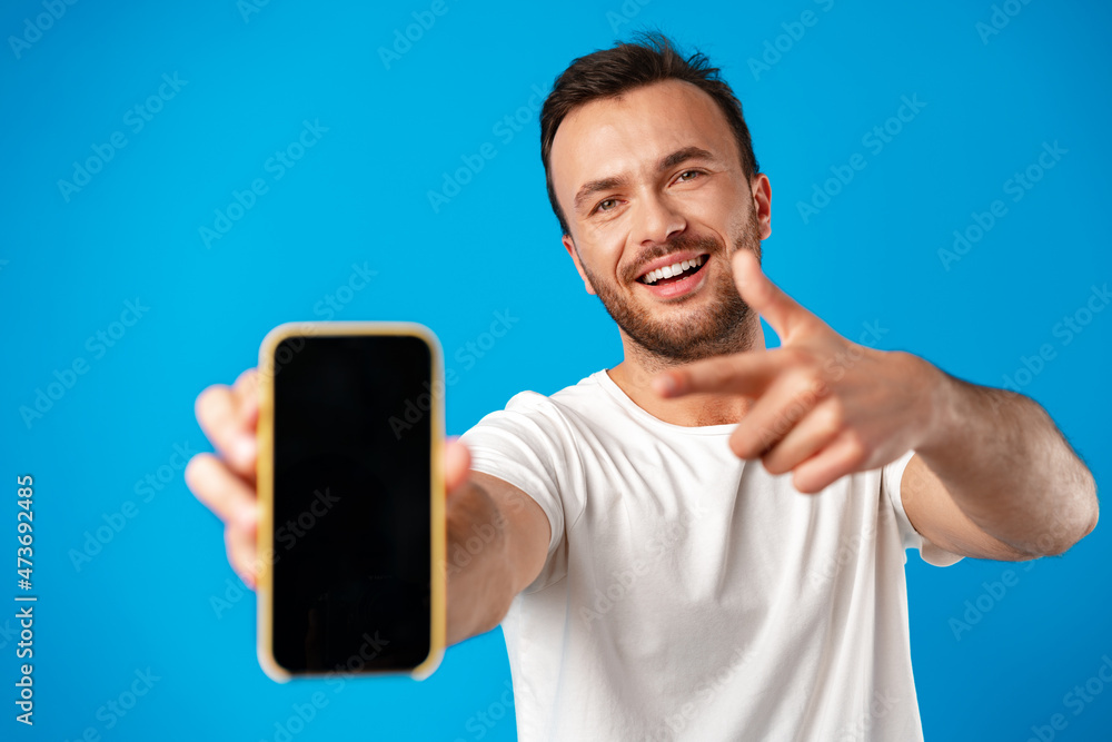 Portrait of young man advertising new smartphone, showing it to camera against blue background