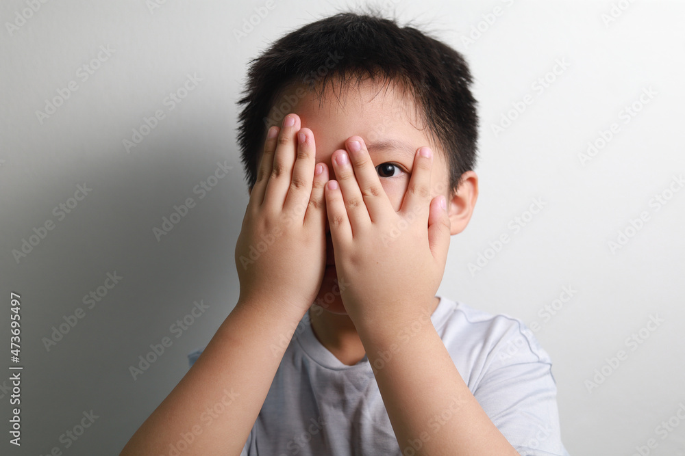 A scared Asian boy covering his eyes with his hands, standing against a white wall. A scared Asian boy covering his eyes with his hands, standing against a white wall.