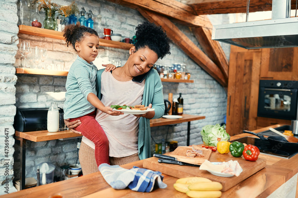 Happy black mother talks on cell phone while preparing breakfast for her daughter in the kitchen. Happy black mother talks on cell phone while preparing breakfast for her daughter in the kitchen.