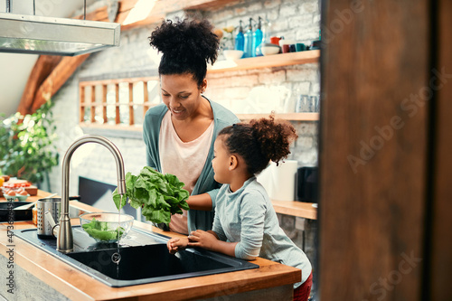 Happy African American mother and daughter wash lettuce under kitchen sink.