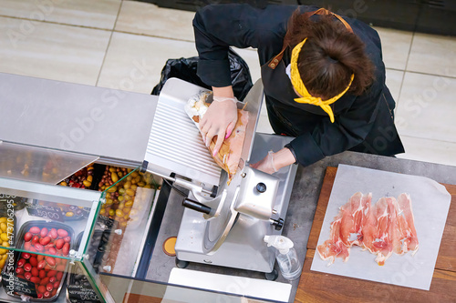 Butcher cut prosciutto, slicing prosciutto on cutting machine. View from above on female butcher cutting meat at butcher counter