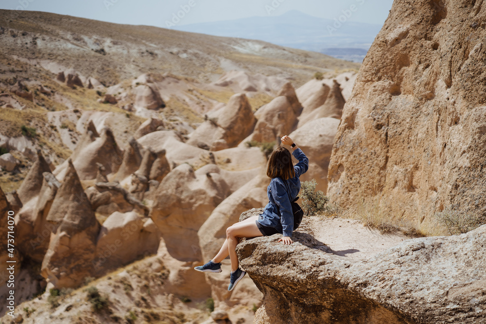 Young happy woman with backpack sitting on a rock with raised hands and looking to the scenary view