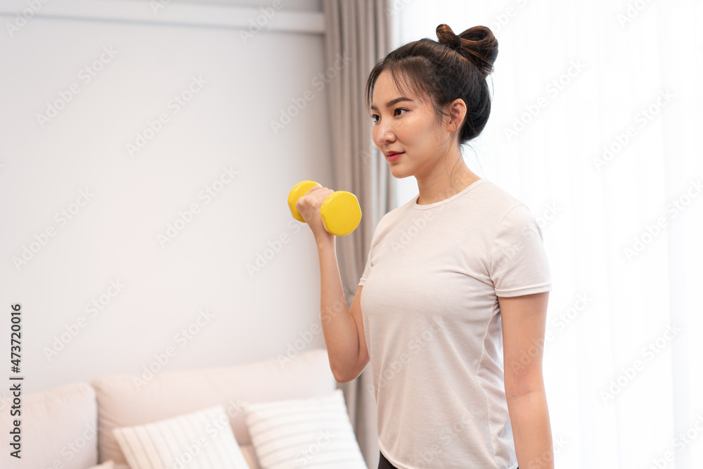 Productive activity concept a girl with a bun wearing a white t-shirt raising the yellow dumbbell with her right hand in her living space Productive activity concept a girl with a bun wearing a white t-shirt raising the yellow dumbbell with her right hand in her living space