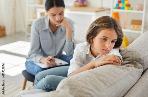 Frustrated little child at psychologist's office. Portrait of girl with sad face sitting on couch during therapy session. Therapist trying to help unhappy, resentful kid who has behaviour issues