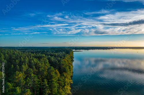 Aerial landscape of the lake and green forest in a sunny day. Blue sky, mirror image of clouds on the water. 