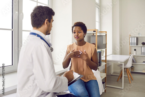 Black woman visiting hospital. Female patient sitting on couch in exam room and talking to professional doctor about chest pain. General practitioner listening to unhappy young lady asking for help