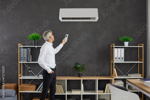 Middle-aged Caucasian man turn on air conditioner in modern home living room. Businessman or CEO switch modern electronic air condition device for fresh ventilated air indoors. Technology concept.