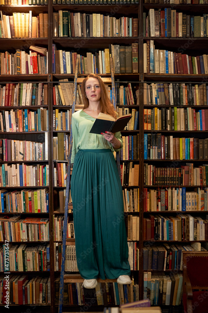 Young woman holding book while standing on ladder in library Young woman holding book while standing on ladder in library