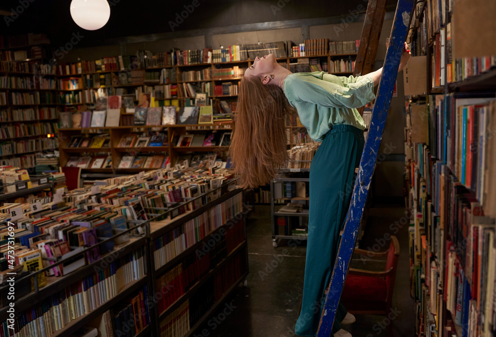 Redhead woman with head back standing on ladder in library Redhead woman with head back standing on ladder in library