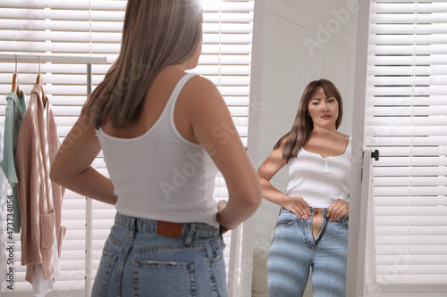 Young woman trying to put on tight jeans near mirror at home photo