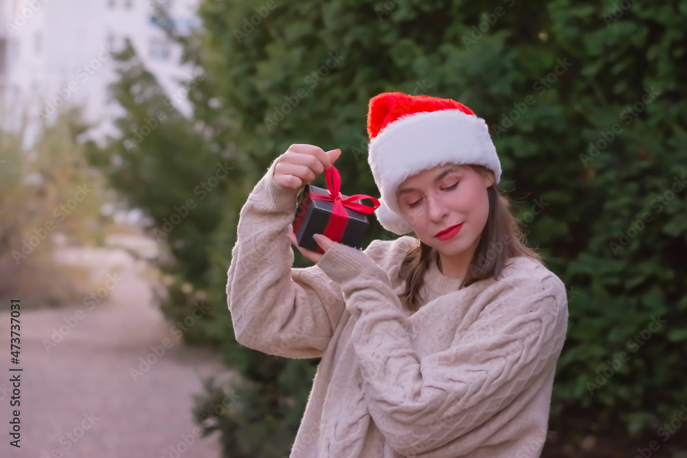 Close up woman in Christmas cap shaking red box at her ear trying to guess what is inside standing