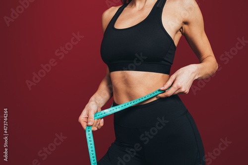 Mid adult sportswoman measuring waist with tape against maroon background photo