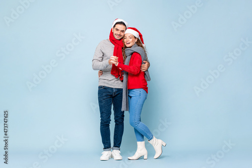 Full length portrait of happy Caucasian couple in Christmas outfits holding each other in light blue isolated studio background