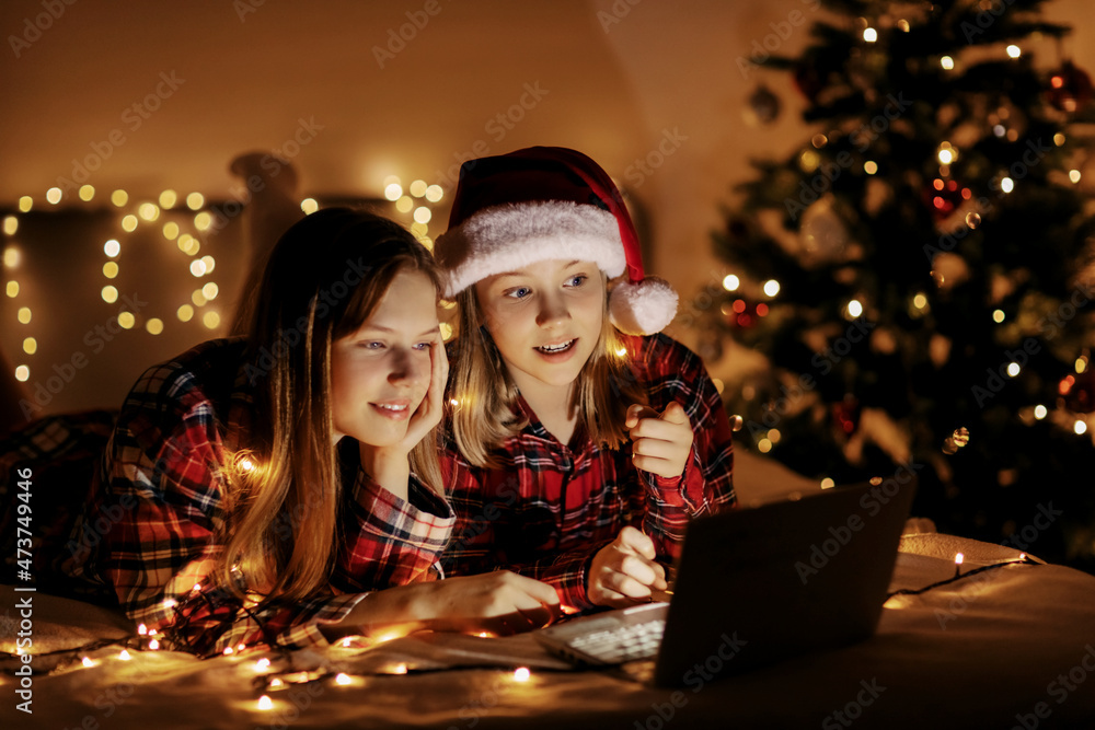 Two charming sisters lie on the bed with a laptop on Christmas or New Year's eve and congratulate their friends and relatives online. Christmas greetings online, virtual party for Christmas Two charming sisters lie on the bed with a laptop on Christmas or New Year's eve and congratulate their friends and relatives online. Christmas greetings online, virtual party for Christmas