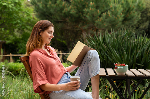 Woman with coffee mug reading book while sitting on chair in garden