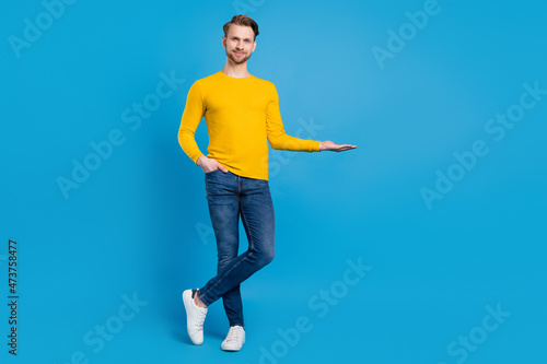 Full length body size of young guy smiling keeping blank space on hand isolated vibrant blue color background