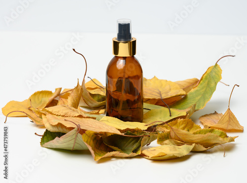 glass brown bottle with cosmetic spray on white background in the middle of yellow leaves