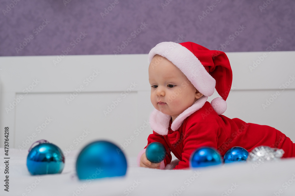 Obraz premium Baby boy dressed as Santa is playing with Christmas decorations on the bed.