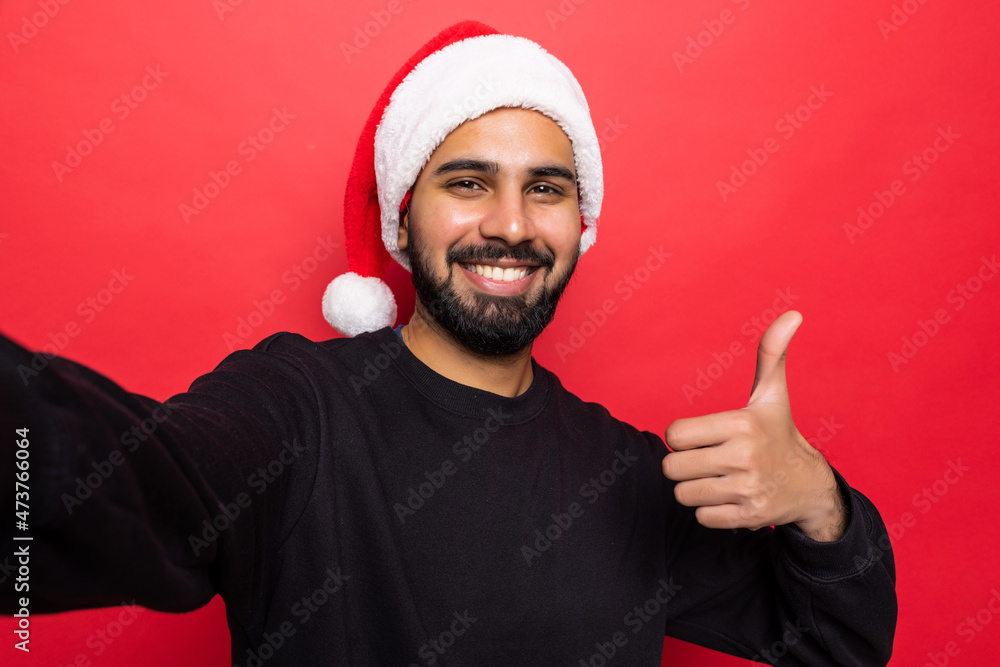 Handsome young man wearing santa hat, taking selfie on mobile phone , standing over red background Handsome young man wearing santa hat, taking selfie on mobile phone , standing over red background