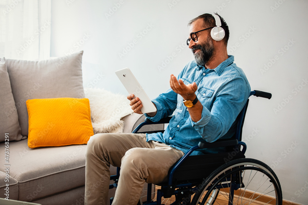 Happy man with headphones and mobile device in wheelchair at home. Photo of mature man in Wheelchairs who is Sitting in the Living Room and Happy man with headphones and mobile device in wheelchair at home. Photo of mature man in Wheelchairs who is Sitting in the Living Room and