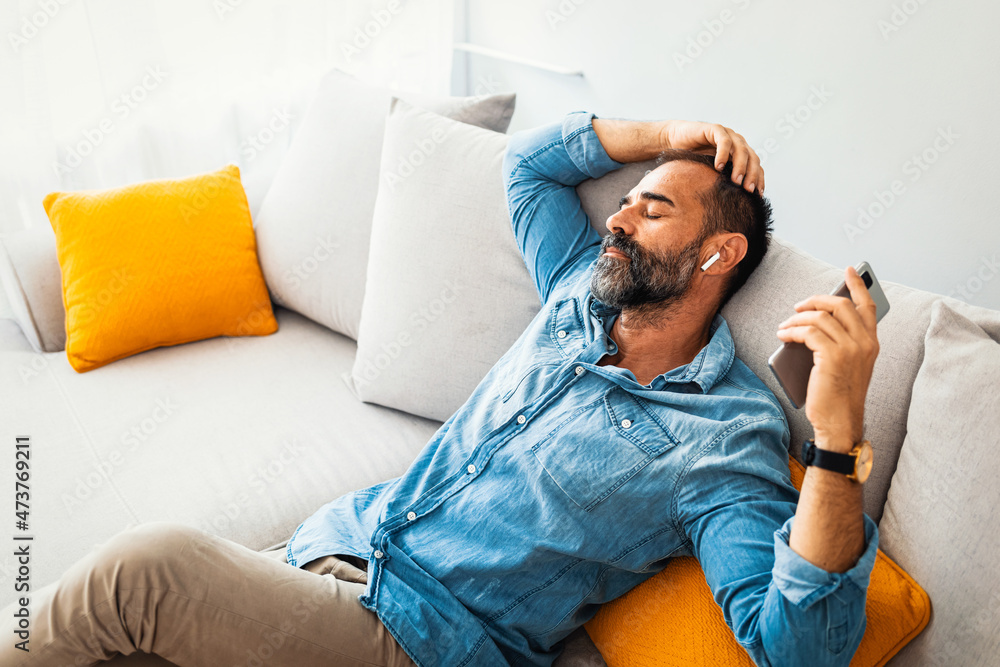 Obraz premium Happy relaxed man with a blissful smile sitting listening to music on his mobile phone using an earbud as he sits on a sofa at home. Man relaxing holding using phone enjoy favorite songs relaxing