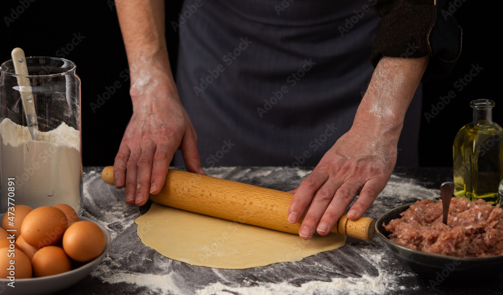A man rolls out the dough with a rolling pin for a russian traditional dish dumplings, ingredients for the dough and minced meat on the table A man rolls out the dough with a rolling pin for a russian traditional dish dumplings, ingredients for the dough and minced meat on the table