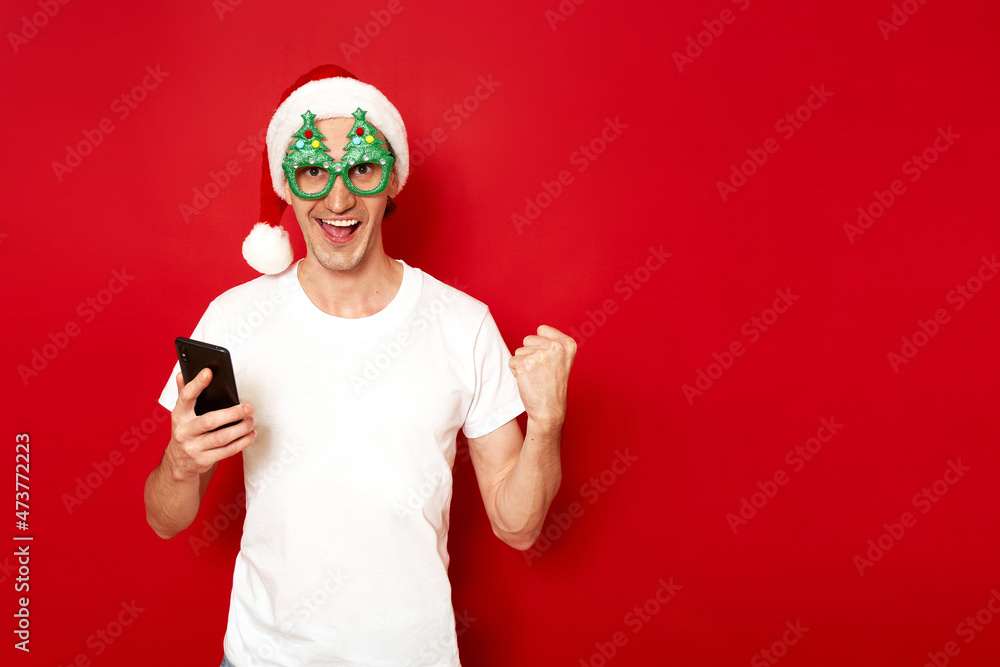 an excited happy man with phone in hands, white T-shirt, Christmas hat, celebrates victory, rejoices in good news notification in application. concept - new year, people, technology, communication