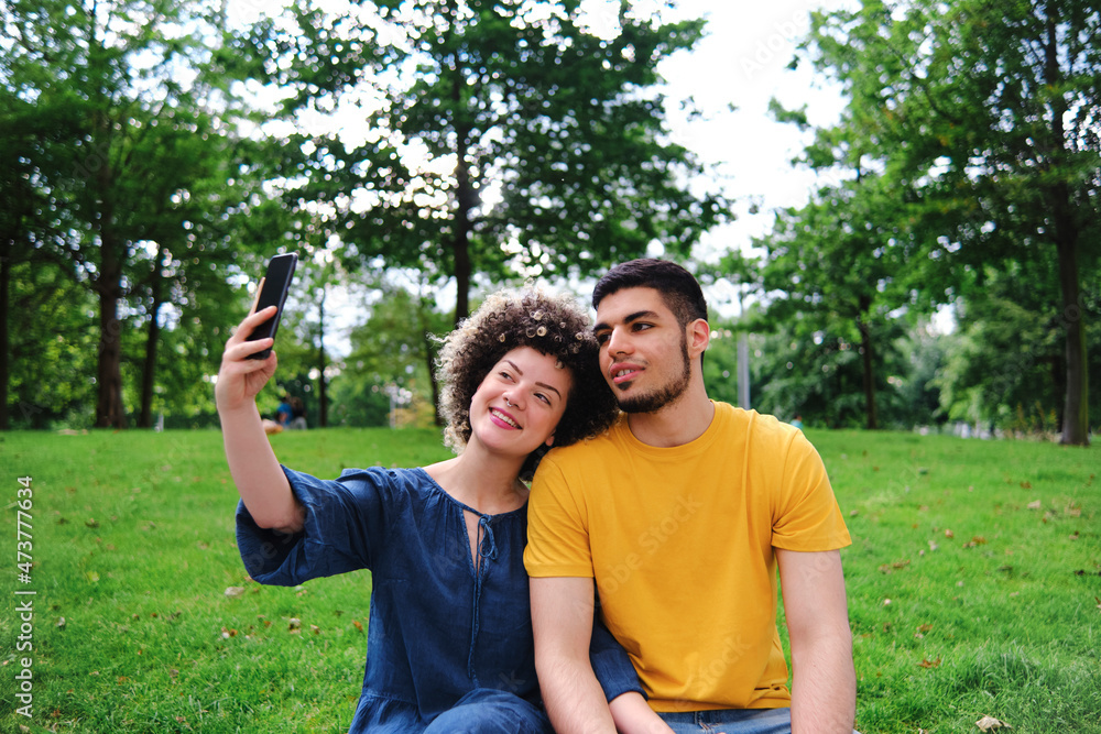 Smiling young woman taking selfie with boyfriend through smart phone while sitting at park Smiling young woman taking selfie with boyfriend through smart phone while sitting at park