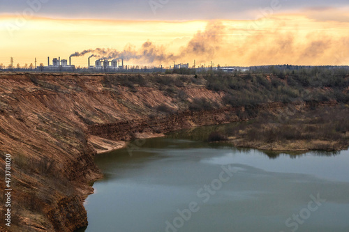 The plant emits smoke at an orange sunset, air pollution through pipes with precise smog against the background of a red quarry with water and overgrown