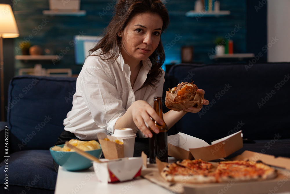 Woman holding beer bottle eating tasty takeaway hamburger while relaxing on couch in front of tv table with fast food menu. Hungry person enjoying junk food sitting on sofa in the evening.