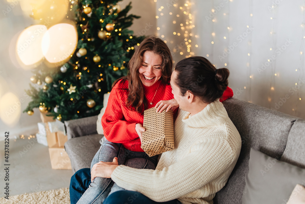 Young couple with gifts in front of Christmas tree. Winter holidays celebration. Giving gifts. Couple in decorated home