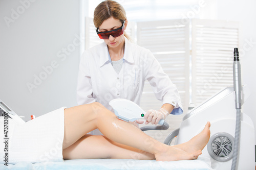 Photo of a cute doctor woman in special protective glasses  who performs a laser hair removal procedure  leg hair removal by modern methods
