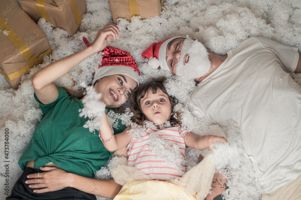 Obraz premium Christmas photo of happy Family. New Year Portrait of dad, mom and daughter wearing Christmas red hats, smiling and laying in white artificial snow. 