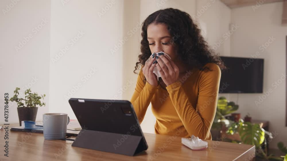 Sick multi-cultural young female blowing her nose while working on electronic tablet in modern home. Sick multi-cultural young female blowing her nose while working on electronic tablet in modern home.