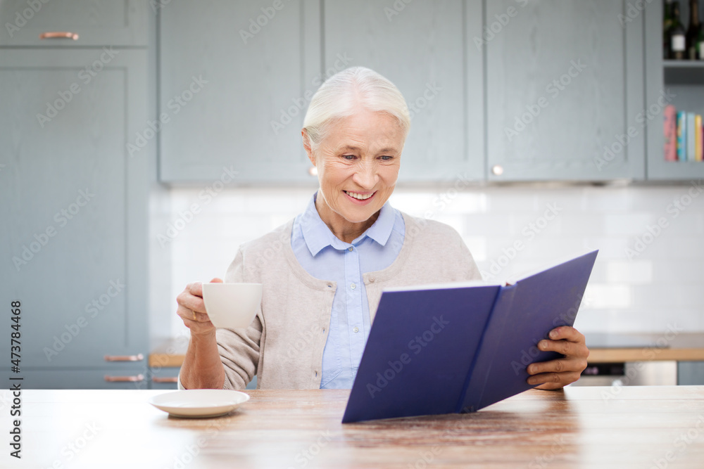 leisure and people concept - happy smiling senior woman drinking coffee and reading book at home over kitchen background leisure and people concept - happy smiling senior woman drinking coffee and reading book at home over kitchen background