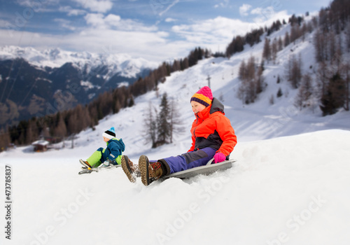 childhood, sledging and season concept - happy little kids sliding on sleds down snow hill in winter over alps mountains on background