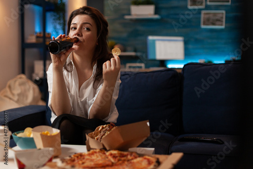 Woman dressed casual drinking beer from bottle while holding a potato chip looking at television comedy sitcom show. Person sitting on couch watching tv in front of table with fast food.