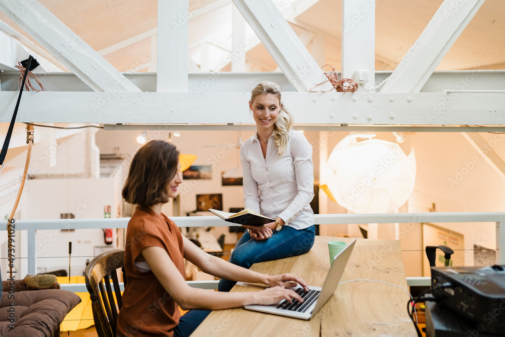 Female entrepreneur looking at colleague typing on laptop sitting at desk in creative office Female entrepreneur looking at colleague typing on laptop sitting at desk in creative office