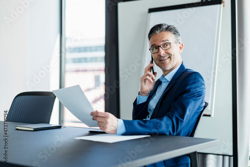 Mature male entrepreneur talking on smart phone sitting at desk looking away photo