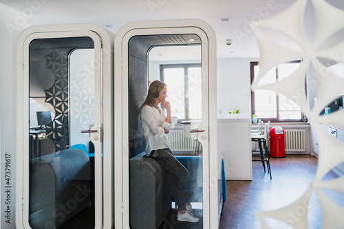 Businesswoman wearing headphones leaning in soundproof cabin at office