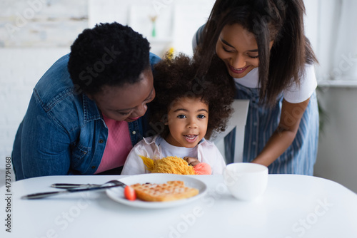 cheerful african american girl sitting at table with blurred breakfast near smiling granny and mom